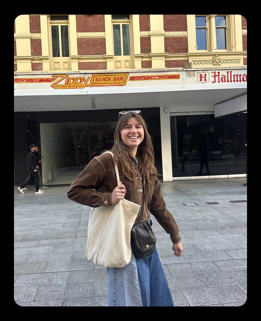 Juliet smiling on a street in Perth, wearing a brown jacket and carrying a tote bag